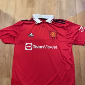 Manchester United tröja 22/23 - Säljer en Manchester United fotbollströja från Adidas med vit krage och svarta detaljer. Tröjan har korta ärmar och klubbmärke på bröstet. Perfekt för fotbollsfans!