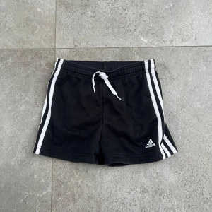 Svarta shorts från Adidas - Snygga svarta shorts från Adidas med vita ränder längs sidorna och en bekväm snörning i midjan. Perfekta för träning eller en avslappnad dag. Adidas-loggan är diskret placerad på ena benet.