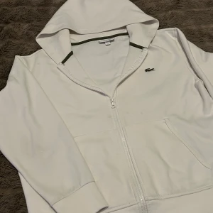 Vit huvtröja från Lacoste - Säljer en stilren vit huvtröja från Lacoste med dragkedja och broderad logga på bröstet. Tröjan har långa ärmar och en bekväm huva med gröna detaljer. Perfekt för en avslappnad look.