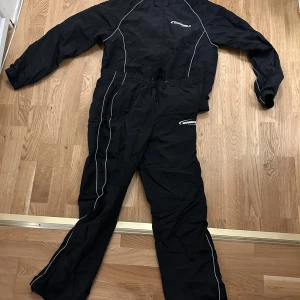 Svart overall från Wuwuco - Säljer en svart overall från Wuwuco med vita detaljer och dragkedja framtill. Overallen har långa ärmar och är perfekt för utomhusaktiviteter. Den har en elastisk midja med snörning för bättre passform.