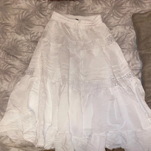 Vit långkjol med spetsdetaljer - Säljer en vacker vit långkjol med spetsdetaljer. Kjolen har en luftig och flödande design med elastisk midja för bekväm passform. Perfekt för varma dagar och ger en romantisk touch till din outfit.