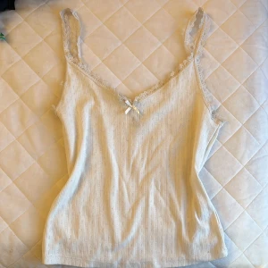 Vit linnetopp från H&M Divided - Säljer en söt vit linnetopp från H&M Divided med spetsdetaljer och en liten rosett framtill. Toppen har smala axelband och ett diskret mönster i tyget. Perfekt för varma dagar eller som ett lager under en tröja.