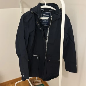 Marinblå regnjacka från Sebago - Säljer en stilren marinblå regnjacka från Sebago. Jackan har en praktisk huva och dragkedja framtill, samt knappar för extra skydd mot väder. Perfekt för regniga dagar med sin klassiska design och funktionella detaljer.
