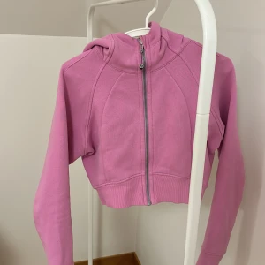Rosa hoodie från Lululemon - Säljer en snygg rosa hoodie från Lululemon med dragkedja framtill. Den har en croppad passform och långa ärmar, perfekt för en avslappnad stil. Hoodien är gjord i ett mjukt material som känns skönt mot huden.