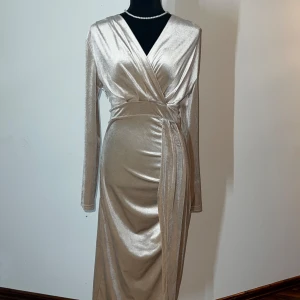 Champagnefärgad omlottklänning - Elegant champagnefärgad omlottklänning med långa ärmar och v-ringad design. Klänningen har en glansig finish och ett knytband i midjan för en smickrande passform. Perfekt för en glamorös kväll.