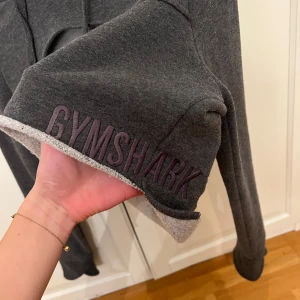 Grå croppad hoodie från Gymshark - Säljer en grå croppad hoodie från Gymshark med en unik design. Den har en öppning framtill och justerbara snören vid huvan. Perfekt för en sportig look eller avslappnad stil. Den är använd 2-3 gånger men tycker inte om stilen på mig. 