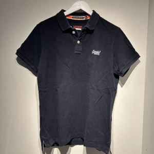 Svart pikétröja från Superdry - Snygg svart pikétröja från Superdry med korta ärmar och klassisk krage. Tröjan har en liten broderad logga på bröstet och knappar framtill. Perfekt för en stilren look.