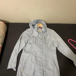 Ljusgrå parkajacka från Simonsen Copenhagen - Snygg ljusgrå parkajacka från Simonsen Copenhagen med dragkedja och knappar framtill. Jackan har en huva och praktiska fickor med dragkedjor. Perfekt för kyligare dagar.