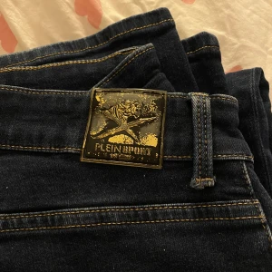 Mörkblå jeans från Plein Sport - Snygga mörkblå jeans från Plein Sport med unika guldfärgade detaljer och ett coolt klösmönster på bakfickan. Jeansen har en klassisk femficksdesign och stängs med dragkedja och knapp. Perfekta för en stilren look.
