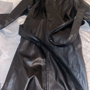 Svart skinnrock med bälte - Snygg svart skinnrock med bälte för en stilren look. Rocken har långa ärmar och knappar framtill. Perfekt för att hålla stilen i kallare väder.