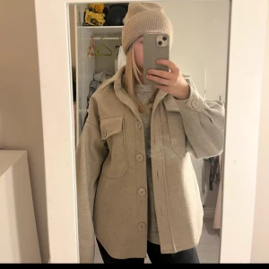 Beige overshirt med knappar - Säljer en stilren beige overshirt med knappar framtill och stora fickor på bröstet. Jackan har en avslappnad passform och är perfekt för lager-på-lager-stil. Den har långa ärmar och en klassisk krage.