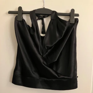 Svart halterneck topp - Snygg svart halterneck topp i satin med knytning i nacken. Toppen har en elegant v-ringning och öppen rygg, perfekt för en stilren look.
