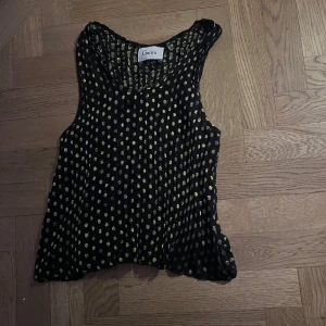 Svart och guldprickig topp från Ganni - Snygg svart topp från Ganni med guldprickigt mönster. Toppen är ärmlös och har en luftig passform, perfekt för en stilren look. Den är tillverkad i ett lätt material som ger en elegant känsla. Står ingen storlek men skulle säga att den är xs/s