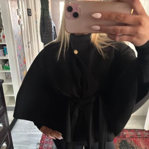 Svart kappa med knytband - Snygg svart kappa med knytband i midjan och dekorativ knappdetalj vid halsen. Perfekt för att ge en elegant touch till din outfit. Passar bra till både vardag och speciella tillfällen.