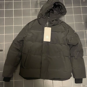 Svart dunjacka från Canada Goose - Säljer en svart dunjacka från Canada Goose med huva och dragkedja. Jackan har långa ärmar och ribbade muddar. Perfekt för kalla dagar. Levereras med originalförpackning.