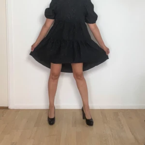 Svart klänning med puffärmar - Charmig svart klänning med puffiga kortärmar och en söt öppen ryggdetalj med knytning. Klänningen har en luftig passform och volangdetaljer som ger en lekfull känsla. Perfekt för att känna sig både bekväm och stilren.