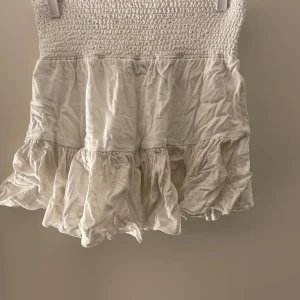 Vit kjol med volangdetaljer - Charmig vit kjol med smockad volang Perfekt för en somrig look med sin luftiga design och korta längd. Kjolen har en söt och romantisk stil som passar perfekt för varma dagar.