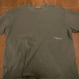 Olivgrön t-shirt från Yoke - Säljer en olivgrön t-shirt från Yoke med texten 'ALTERED CLUB' tryckt på bröstet. T-shirten har en klassisk rund hals och korta ärmar. Perfekt för en avslappnad stil. Köpt på Junk yard