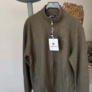 Olivgrön tröja från Boss - Snygg olivgrön tröja från Boss med dragkedja och ribbad krage. Perfekt för en stilren look. Passar bra till både jeans och chinos. På verkligheten ser det finare ut än bilder