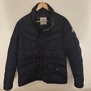  Moncler jacka - Säljer en stilren svart dunjacka från Moncler med broderad logga på ärmen. Perfekt för våren. Jackan har långa ärmar och flera praktiska fickor framtill. Perfekt för kyliga dagar med sin quiltade design och knappar för extra stil. Jackan är av en äldre modell därför så finns det ingen qr kod utan en vanlig kod. Skriv för fler bilder och vid minsta intresse