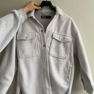Ljusgrå overshirt från Zara - Säljer en stilren ljusgrå overshirt från Zara med knappar framtill och två stora bröstfickor. Overshirten är något längre baktill. 