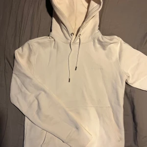 Vit hoodie från J.Lindeberg - Säljer en stilren vit hoodie från J.Lindeberg i storlek S. Den har en klassisk design med dragsko i huvan och en stor ficka framtill. Perfekt för en avslappnad look.