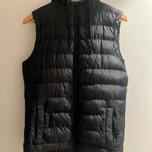 Svart dunväst med huva - Snygg svart dunväst med dragkedja och huva. Perfekt för kyligare dagar när du vill hålla värmen utan att bära en jacka. Västen har en quiltad design och praktiska fickor framtill.