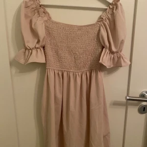 Beige klänning med puffärmar - Säljer en söt beige klänning med puffiga kortärmar och smockdetaljer på överdelen. Perfekt för en somrig look. Klänningen har en luftig passform och är gjord i ett lätt material.