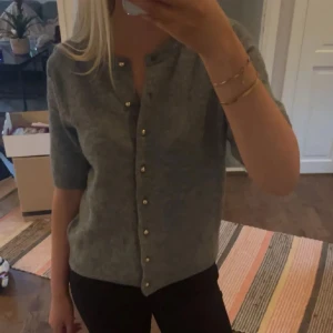 Grå kofta med knappar - Säljer en stilren grå kofta med korta ärmar och knappar framtill. Perfekt för lager-på-lager och passar till många olika outfits. Koftan är mjuk och bekväm, idealisk för både vardag och lite finare tillfällen.