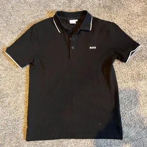 Svart kortärmad pikétröja från Boss - Säljer en stilren svart pikétröja från Boss med vita detaljer på krage och ärmslut. Tröjan har en klassisk passform och är perfekt för en avslappnad men ändå snygg look. Den är kortärmad och har en knappslå framtill.