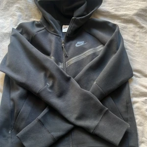 Svart Nike Tech - Säljer en svart huvtröja från Nike med dragkedja och huva. Tröjan har långa ärmar och en diskret Nike-logga på bröstet. Perfekt för en sportig look eller avslappnade dagar.