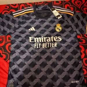 Real Madrid fotbollströja - Snygg Real Madrid fotbollströja från Adidas i mörkblått med ett vågigt mönster. Tröjan har korta ärmar med gulddetaljer och klubbens emblem på bröstet. Perfekt för fans som vill visa sitt stöd med stil.