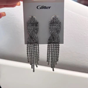 Glittriga örhängen från Glitter - Snygga glittriga örhängen från Glitter med hängande fransar. Perfekta för att ge en extra touch till din outfit. De är i en elegant silverfärg och har en iögonfallande design. Helt nya.