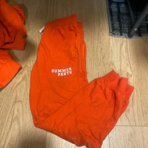 Orange hoodie och byxor - Säljer en cool orange hoodie med texten 'GAME PICNIC' och ett chipsmotiv på bröstet. Baksidan har ett större tryck med texten 'SUMMER PARTY GAME PICNIC' och 'PLAY WITH FRIENDS BE HAPPY'. Perfekt för avslappnade tillfällen. Plus byxorna som ni ser på första bilden. Det är ett sett för unga killar. 