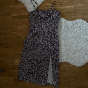 Blommig klänning med slits - Säljer en söt blommig klänning med smala axelband och en stilren slits framtill. Klänningen har en svart bas med små rosa och vita blommor. Perfekt för sommarens alla tillfällen! 🌸