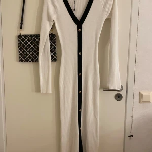 Vit ribbad klänning från Missguided - Snygg vit ribbad klänning från Missguided med svarta detaljer och knappar längs hela framsidan. Klänningen har en v-ringad design och långa ärmar, perfekt för en stilren look.