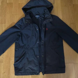 Svart jacka från Polo Ralph Lauren - Säljer en stilren svart jacka från Polo Ralph Lauren med huva och dragkedja. Jackan har en röd broderad logotyp på bröstet och knappar framtill. Perfekt för höst och vår.