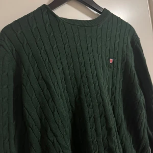 Mörkgrön stickad tröja med kabelmönster  - Säljer en snygg mörkgrön stickad tröja med kabelmönster. Tröjan har en rund halsringning och ett litet emblem på bröstet. Perfekt för kyligare dagar!