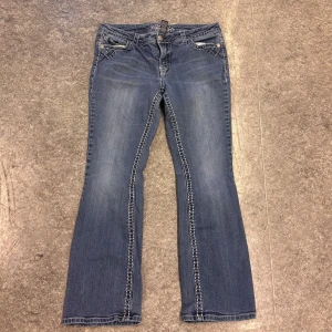 Blå bootcut jeans - Säljer ett par snygga blå bootcut jeans med dekorativa bakfickor. Jeansen har en klassisk femficksdesign och är tillverkade i en bomullsblandning med lite stretch för extra komfort.
