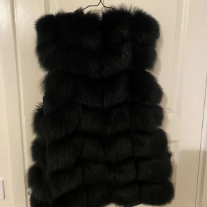 Svart pälsväst - Snygg svart pälsväst som ger en lyxig känsla. Perfekt för att hålla sig varm och stilren under kyliga dagar. Västen har en mjuk insida och en fluffig utsida som ger extra volym. Äkta päls!