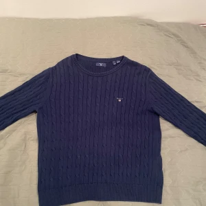 Mörkblå stickad tröja från GANT - Säljer en stilren mörkblå stickad tröja från GANT. Tröjan har ett klassiskt kabelstickat mönster och rund halsringning. Perfekt för kyligare dagar.