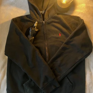 Svart zip hoodie Ralph Lauren - Säljer en klassisk svart hoodie från Polo Ralph Lauren med dragkedja och röd logotyp på bröstet. Perfekt för en avslappnad stil. Hoodien har en mjuk insida och praktiska fickor framtill. Perfekt nu till vintern. Skick 10/10 Inga defekter. Storlek XL i barnstorlek men passar mer som S i vanlig storlek