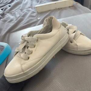 Vita sneakers från Gant - Snygga vita sneakers från Gant med snörning och guldfärgade öljetter. Perfekta för en stilren look. Skorna har en diskret logga på hälen och en bekväm sula.