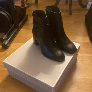 Svarta boots från Tamaris - Snygga svarta boots från Tamaris i skinn med dragkedja och dekorativa remmar. Perfekta för höst och vinter med en bekväm klack och spetsig tå. Har använt en gång 