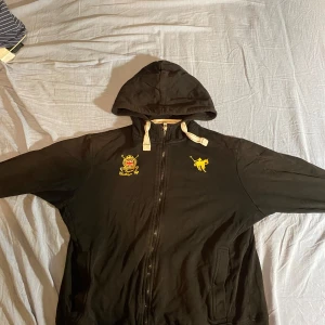 Polo zip hoodie - Säljer en svart huvtröja från polo med dragkedja och broderade detaljer i gult. Tröjan har en justerbar huva med snören och långa ärmar. Perfekt för en avslappnad stil. Skick 8/10 Står strlk M men sitter som S. Skriv till mig om ni har några frågor!