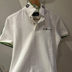 Vit pikétröja från Sail Racing - Snygg vit pikétröja från Sail Racing med gröna och gula detaljer. Tröjan har korta ärmar och en broderad logga på bröstet. På baksidan finns texten 'BRA 77' och 'Sail Racing International'. Perfekt för en sportig look!