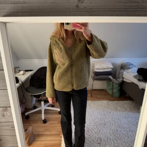 Grön teddyjacka med huva - Mysig grön teddyjacka med dragkedja och huva. Perfekt för kyliga dagar. Jackan har en normal passform och en praktisk bröstficka.