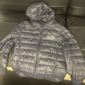 Mörkblå pufferjacka från EA7 - Säljer en mörkblå pufferjacka från EA7 med huva och dragkedja. Jackan är quiltad och har en stilren design med logotyp på bröstet. Perfekt för kyligare dagar.