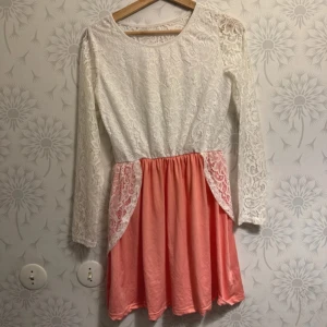 Vit och rosa spetsklänning - Charmig klänning med vit spetsöverdel och rosa kjol. Klänningen har långa ärmar och en vacker spetsdetalj som ger en romantisk känsla. Perfekt för speciella tillfällen.