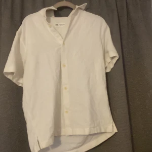 Vit kortärmad skjorta från Zara - Säljer en stilren vit kortärmad skjorta från Zara. Skjortan har en klassisk krage och knappar framtill. Perfekt för varma dagar eller sommarens festligheter.